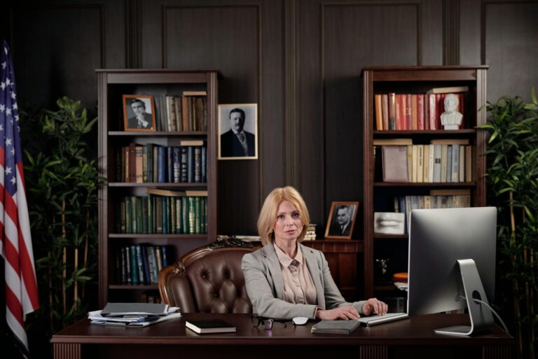 A businesswoman sits confidently at her desk in an office setting with a bookshelf and American flag.